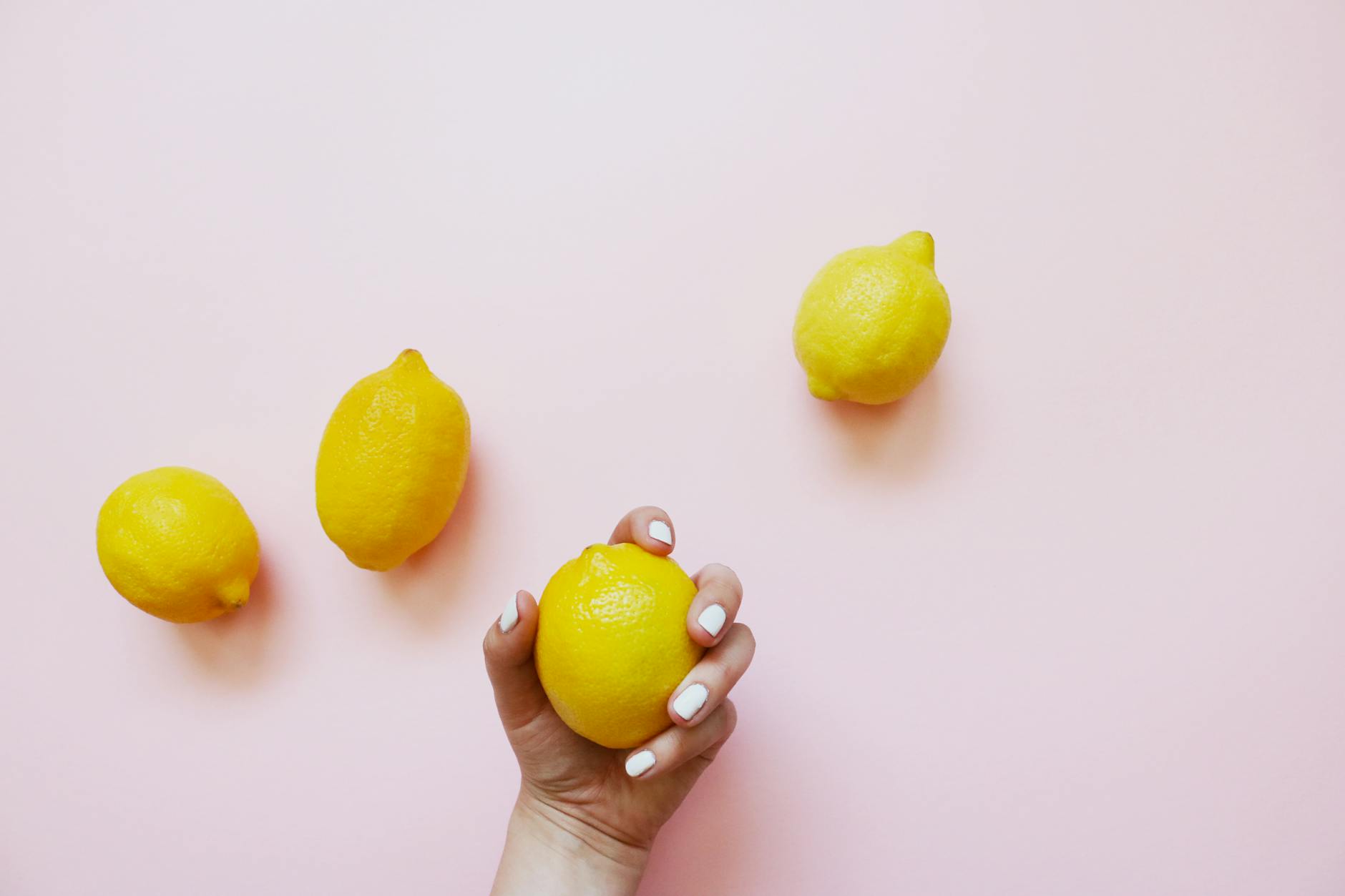 A hand holding a lemon against a soft pink background surrounded by lemons